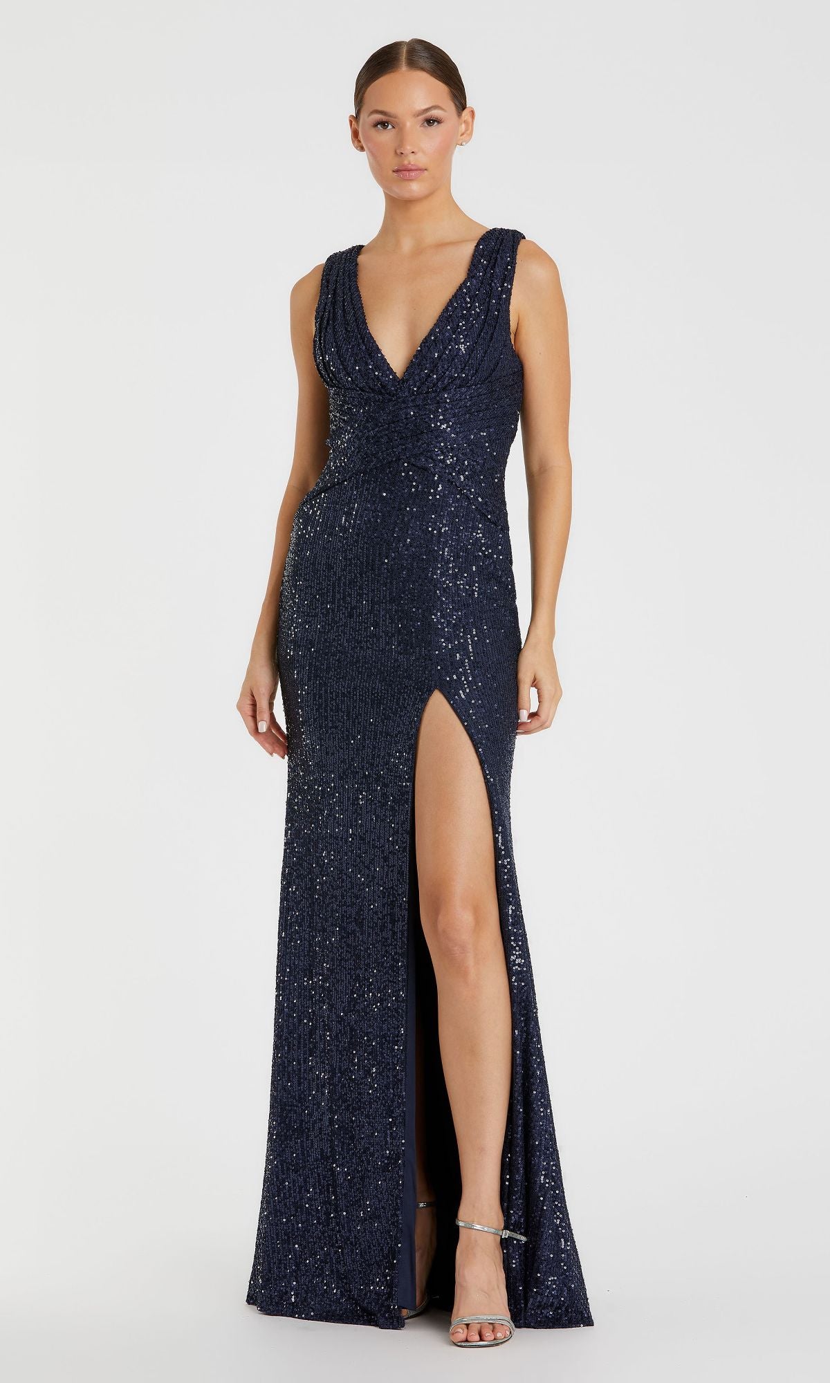 A woman wearing the Mac Duggal 26598 Sleeveless Long Formal Dress, featuring a deep V-neckline, high front slit, dark blue sequins, stands against a plain white background.